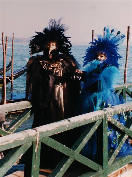 Two Venetian carnival masks in black and blue feathers on a wooden pier, lagoon behind