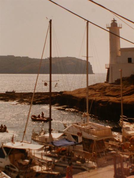Sailboats moored beneath the white lighthouse of Ventotene, sparkling sea and rocks