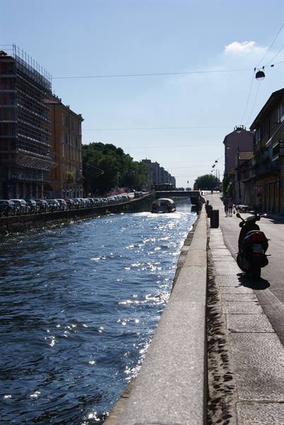 Scooter parked along Milan's Naviglio Grande canal, boat passing, bright sunlight sparkling on water