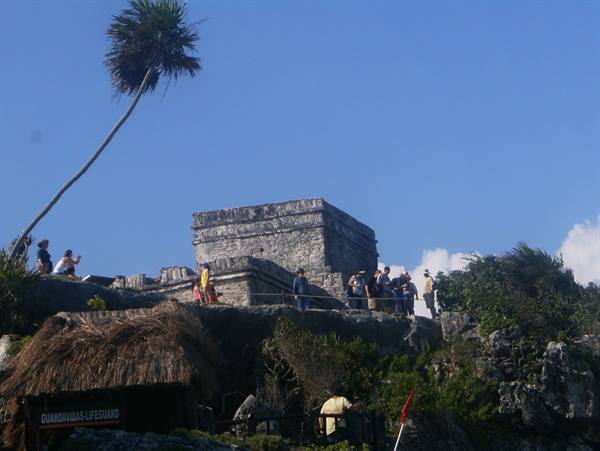 Tourists on Tulum Mayan stone ruins under blue sky, leaning palm