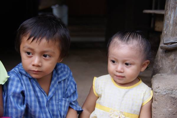 Two children sitting in the shade of a house, one in blue shirt
