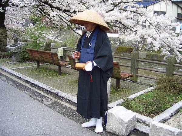 Monk in straw hat collecting alms under cherry blossoms by canal