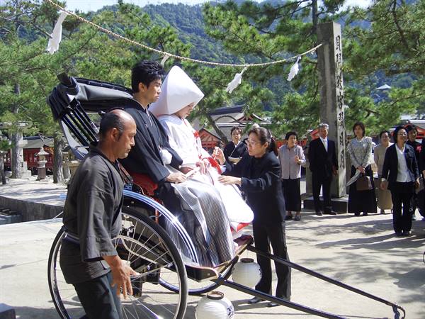 Bride in white kimono on a rickshaw at Itsukushima shrine, guests watching nearby