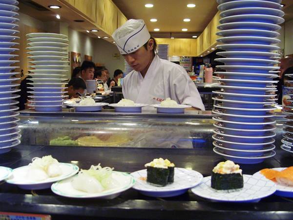 Chef in white prepares sushi among stacked plates at conveyor restaurant