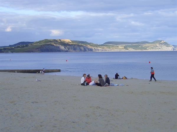 Group sitting on sand before green cliffs across the bay, cloudy sky