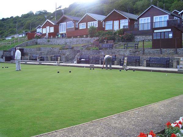 Two lawn bowls players on the green, wooden chalets and benches behind