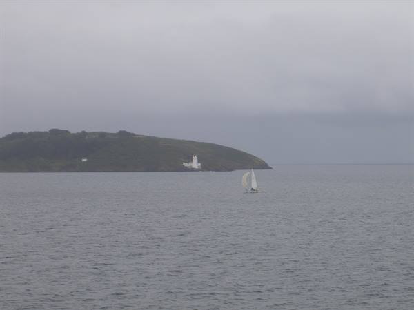 Sailboat on grey sea in front of green headland with white lighthouse