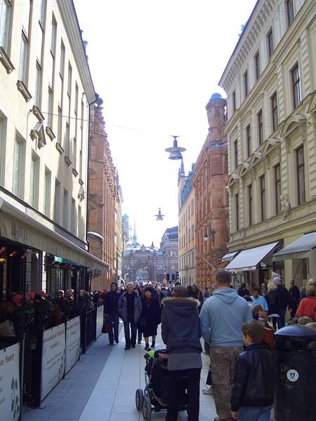 Crowded pedestrian shopping street with brick buildings, café awnings, daylight