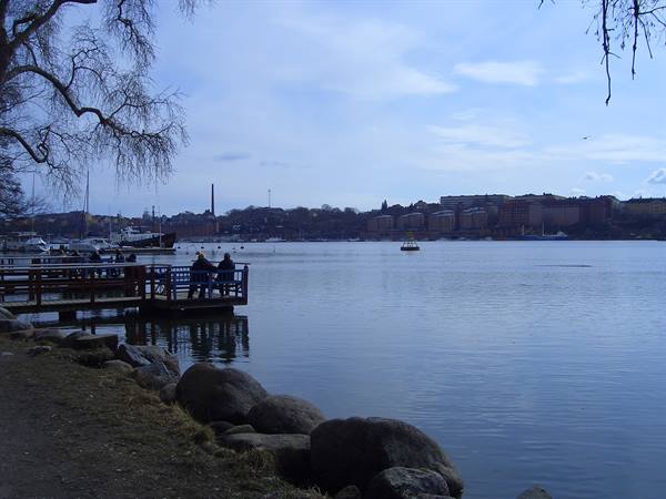Couple on wooden pier by rocks, boats and buildings across calm water, Stockholm
