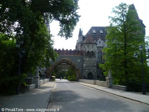 Vajdahunyad Castle: stone bridge with arch, Gothic towers among green trees, daylight