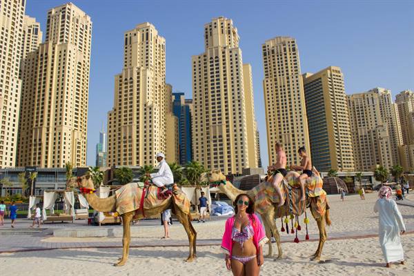 Tourists riding a camel on the beach among Dubai Marina skyscrapers