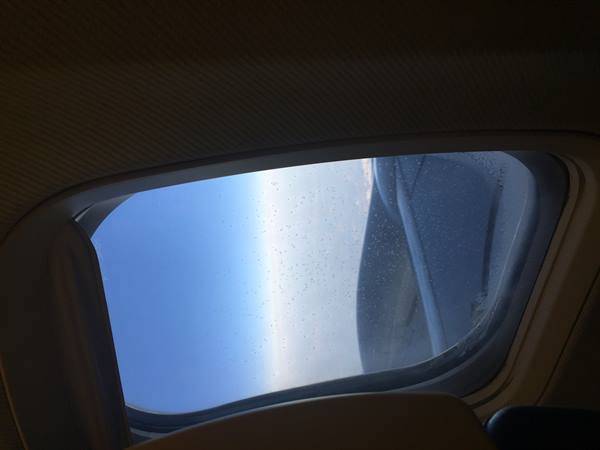 Airplane window with water droplets and wet wing under blue sky