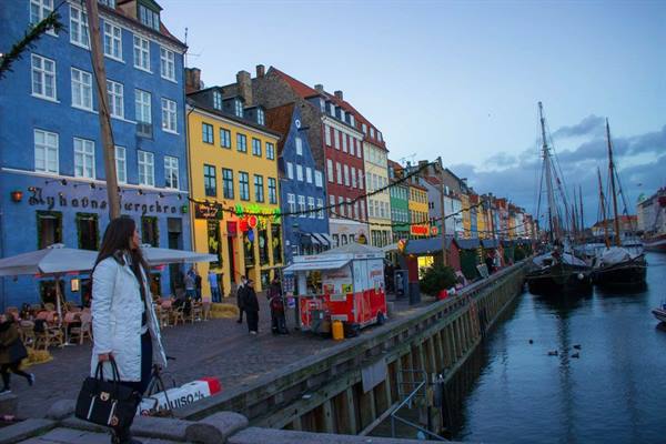 Woman in white coat walks past Nyhavn's colorful townhouses with moored boats