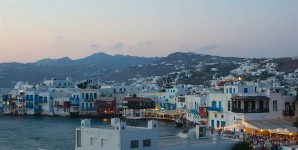 White houses along Mykonos waterfront, lights glowing, mountains at dusk