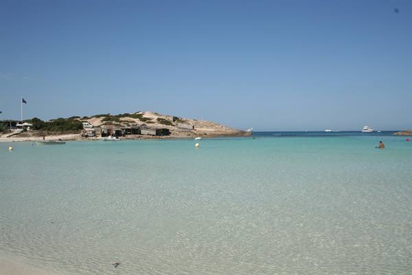 Clear turquoise water over sandy beach, rocks and boats under blue sky