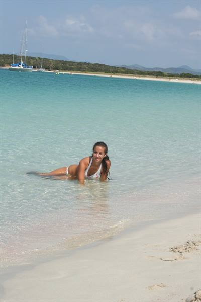 Woman in bikini lying in clear turquoise water, sailboat and white beach behind
