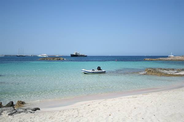 Inflatable boat anchored in clear turquoise water, white sand beach, ships on horizon