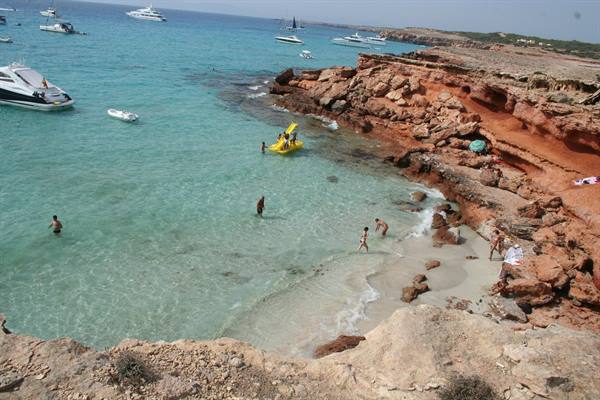 Swimmers and yellow dinghies among red cliffs and turquoise water at Cala Saona