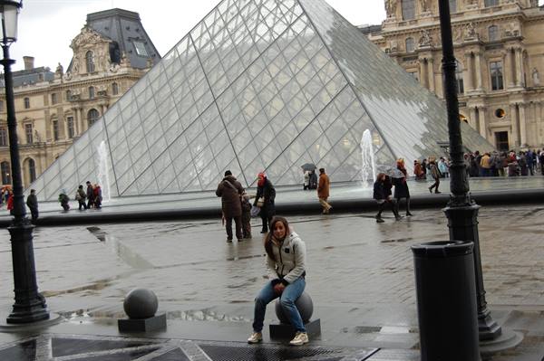 Girl sitting on a stone sphere in front of the Louvre glass pyramid