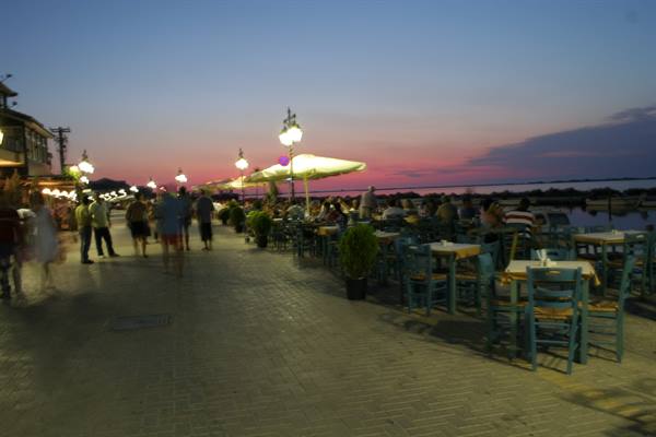 Outdoor tables with blue chairs along the waterfront, people strolling under lit lampposts