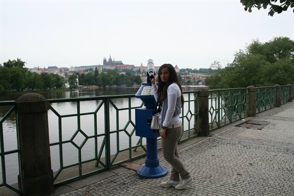 Tourist next to a blue coin-operated binocular on the Vltava riverbank, Prague Castle behind