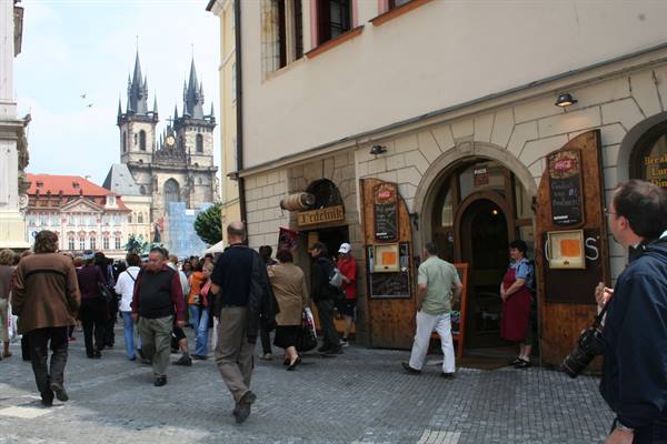 Tourists crowd a cobbled street outside a pub, with Týn Church looming in Prague
