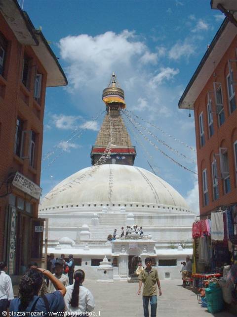 The great Boudhanath stupa among brick houses, people strolling by, prayer flags