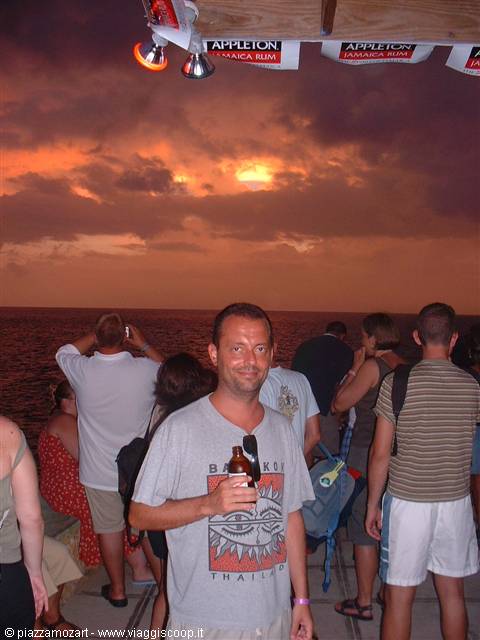 Tourist holding beer on boat deck, orange clouds glowing over the sea