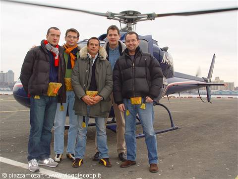 Six men in yellow flight suits pose in front of a blue helicopter on a helipad