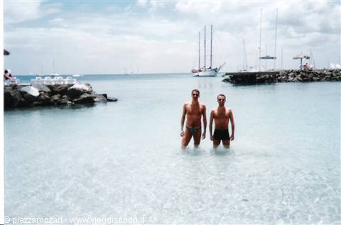 Two men in swim trunks in turquoise water near sailboats and rocky breakwater