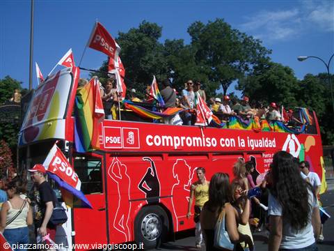 Red double-decker PSOE bus with rainbow flags and people on top, Madrid