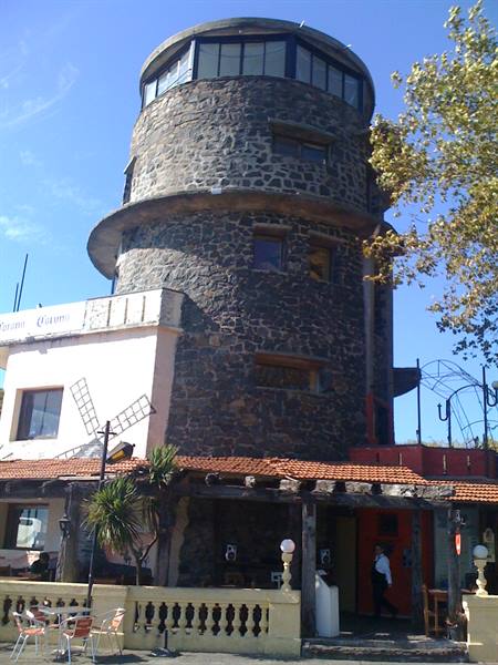 Volcanic stone tower with rooftop glass windows, outdoor restaurant tables below, sunshine