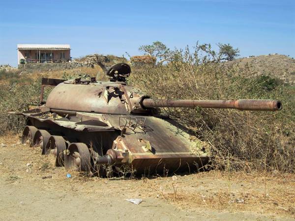 Rusted tank abandoned among dry bushes, white house in the background