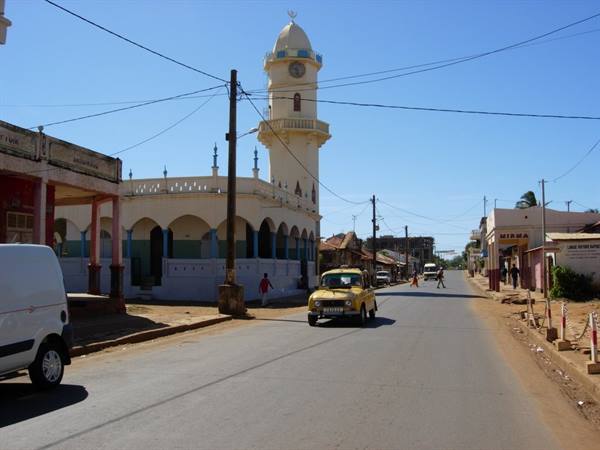Yellow car on dirt road in front of a white mosque with minaret under blue sky