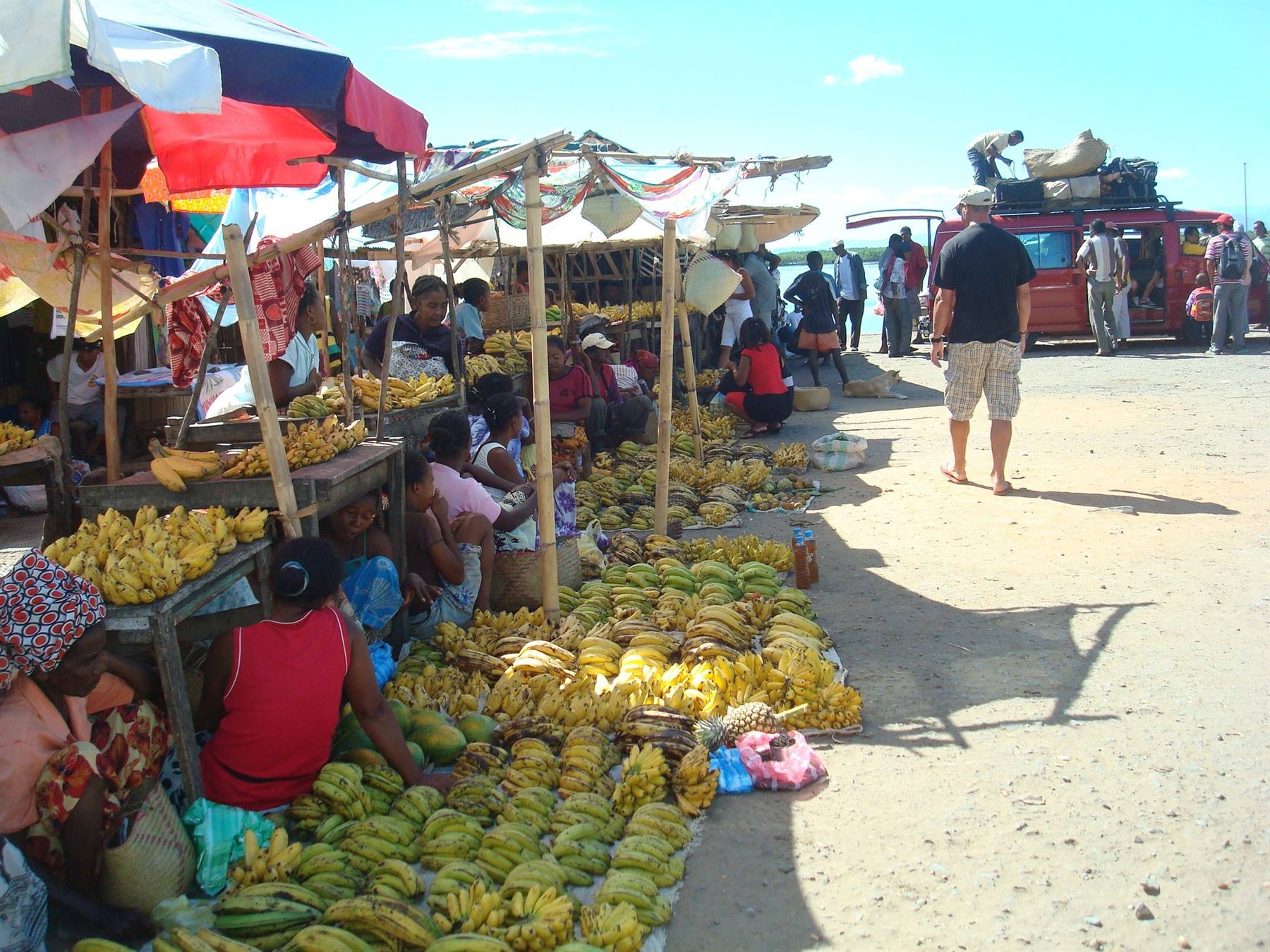 Mercato di banane su sabbia, venditrici sotto ombrelloni, turista cammina vicino un furgone
