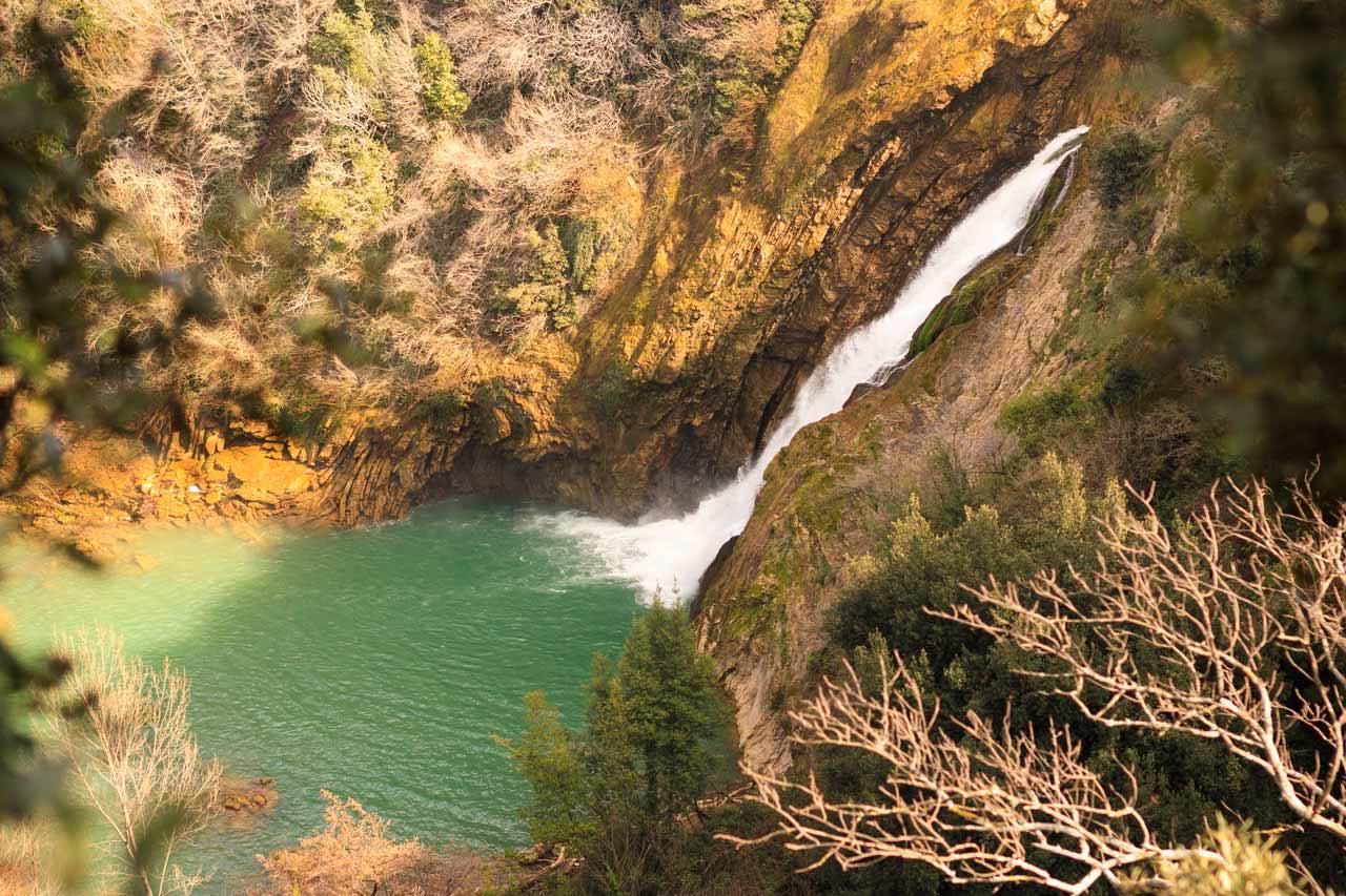 Cascata tra rocce rade in una pozza verde smeraldo, rami spogli