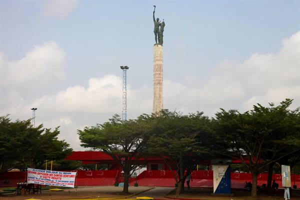 Tall column topped by bronze statue above red stands and trees