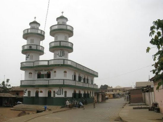 Moschea bianca e verde con due minareti a terrazze, strada di terra con passanti