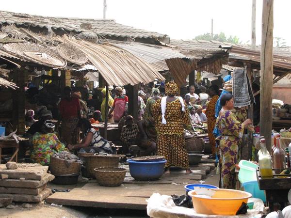 Women in colorful dresses sell goods at a thatched-roof market