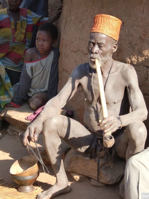 Anziano con cappello arancione e copricapo di paglia fuma una lunga pipà di legno accanto a un bambino, davanti a case in fango