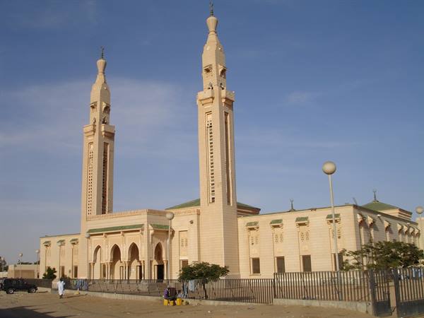 Light stone mosque with two tall minarets, blue sky, small market below