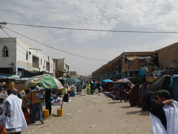 Sandy street market, green umbrellas, stalls, low buildings, people in white robes