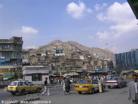 Yellow taxis and pedestrians cross a busy street beneath hillside houses in Kabul