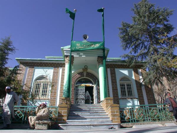 Brick mosque with green flags, men sitting on stairs under blue sky
