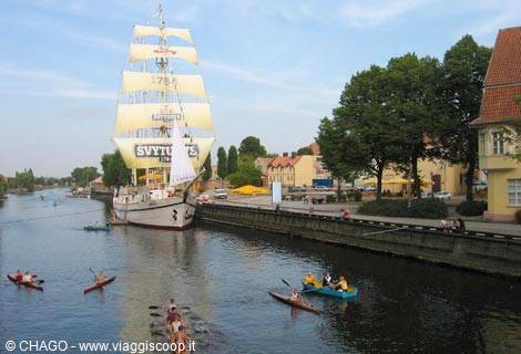 White-sailed tall ship moored along the canal, colorful kayaks paddling nearby