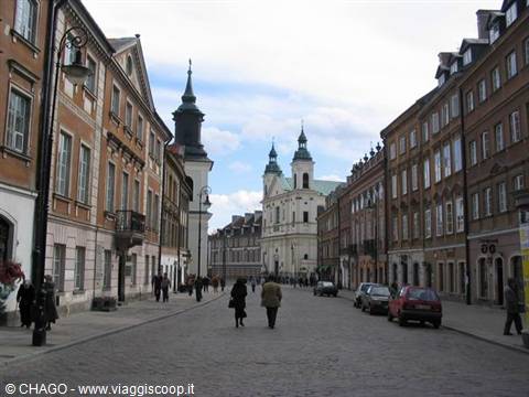 Pedestrians on a cobbled street past historic buildings and white twin-towered church in Warsaw