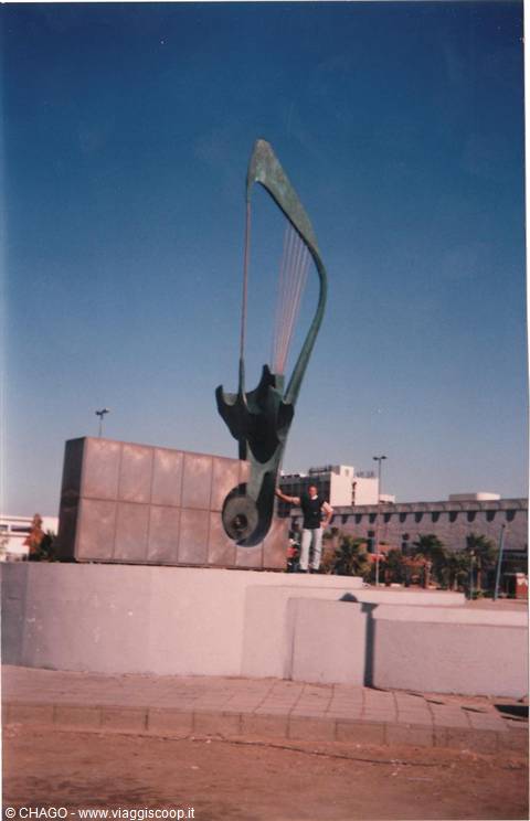 Man posing beside a large metal harp sculpture on a stone base