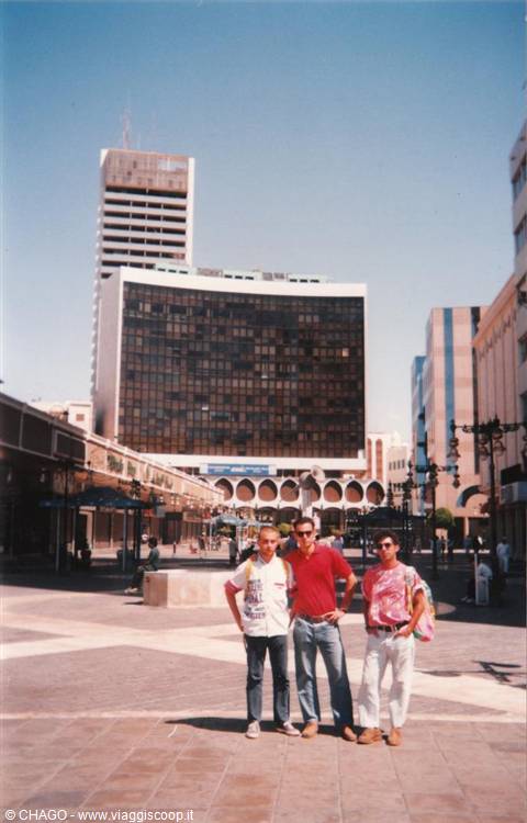 Three tourists pose on a paved square with a skyscraper behind