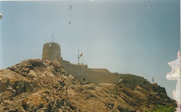 Stone fort with round tower on rocky hill, flags waving, Muscat
