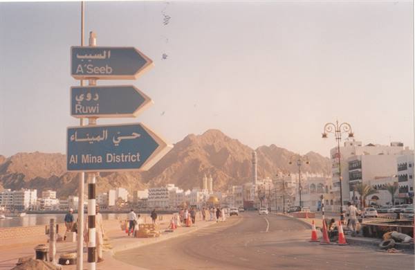 Blue road sign pointing to A'Seeb and Al Mina District, arid mountains behind, Muscat
